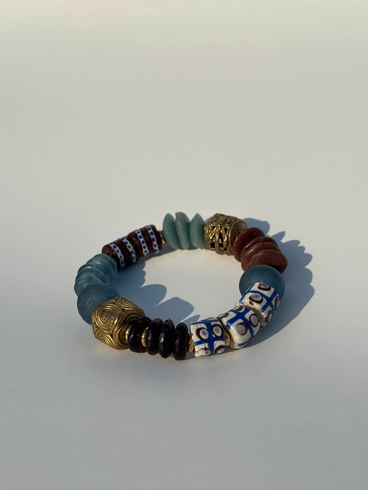 A beaded stretch bracelet featuring a mix of matte blue-gray, metallic gold, brown, white, and blue patterned beads, displayed on a light gray background.