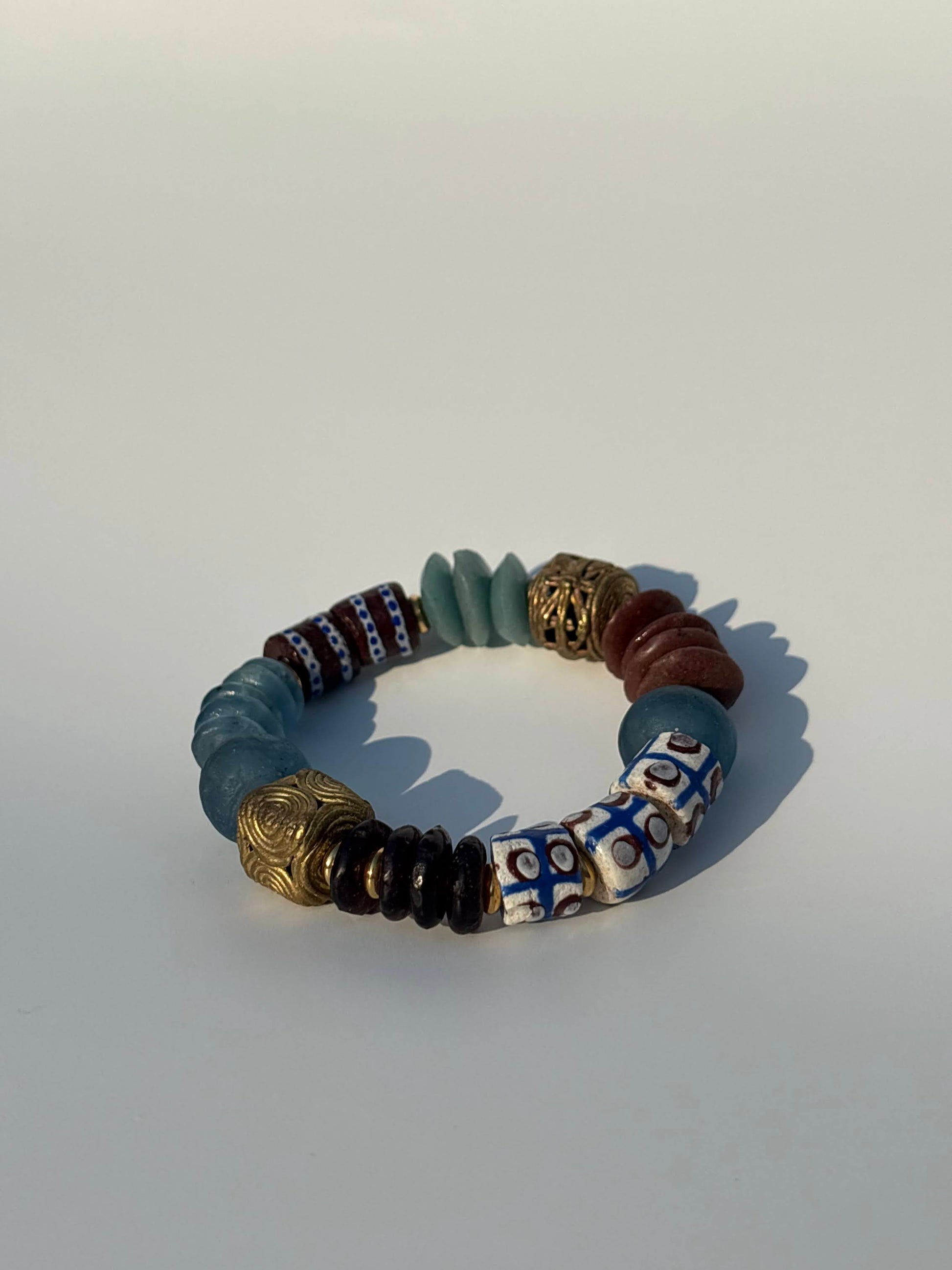 A beaded stretch bracelet featuring a mix of matte blue-gray, metallic gold, brown, white, and blue patterned beads, displayed on a light gray background.