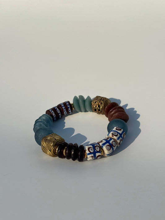 A beaded stretch bracelet featuring a mix of matte blue-gray, metallic gold, brown, white, and blue patterned beads, displayed on a light gray background.